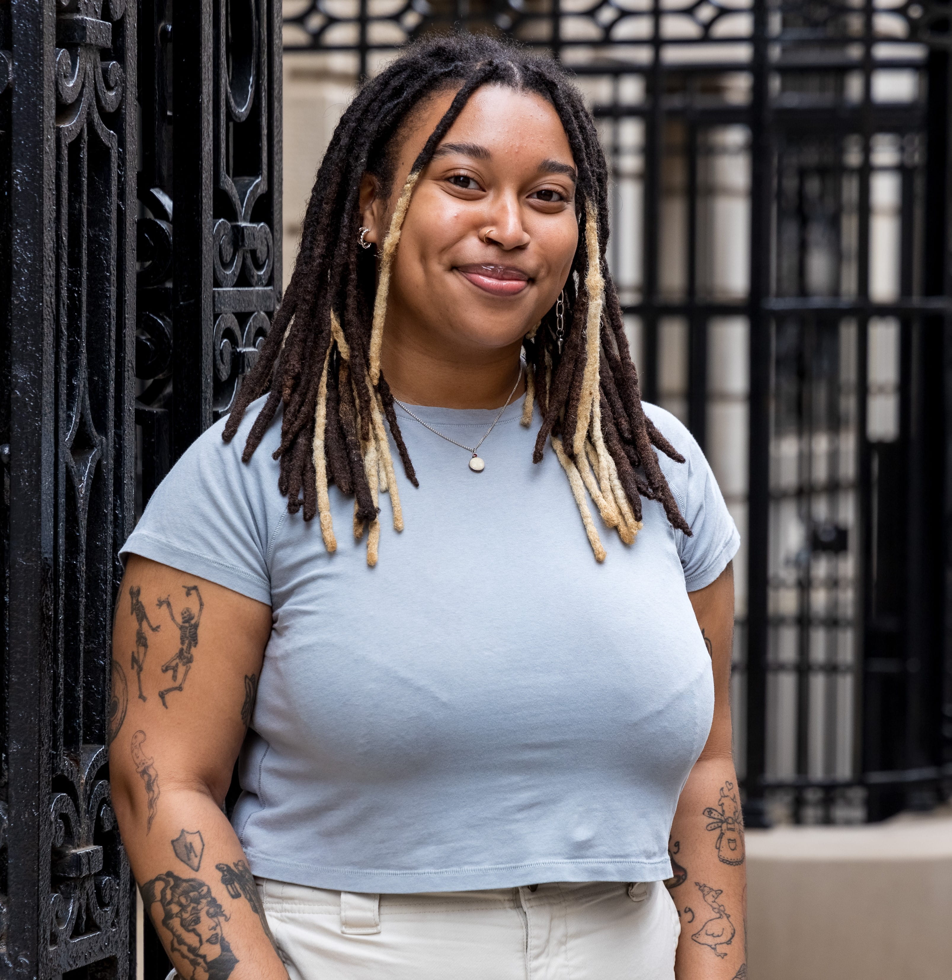 Half-length portrait of a smiling young person with dreadlocks, wearing a light blue top in front of a gate in NYC